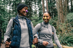 Mid-range shot of two women hiking in the woods with arms intertwined. Both women are sporting Nöz sunscreen on their nose as they smile at each other.